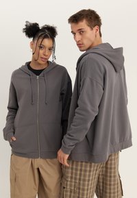 Felpa grigia con zip, cappuccio, tasche frontali e cordino, caratterizzata da una vestibilità comoda. I modelli indossano pantaloni cargo beige con un motivo a quadri.