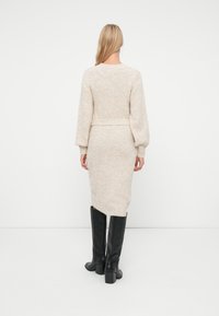 Vestito lavorato a maglia beige con lunghe maniche a sbuffo, una cintura in vita e un orlo leggermente curvo sul retro. Indossato con stivali neri fino al ginocchio.