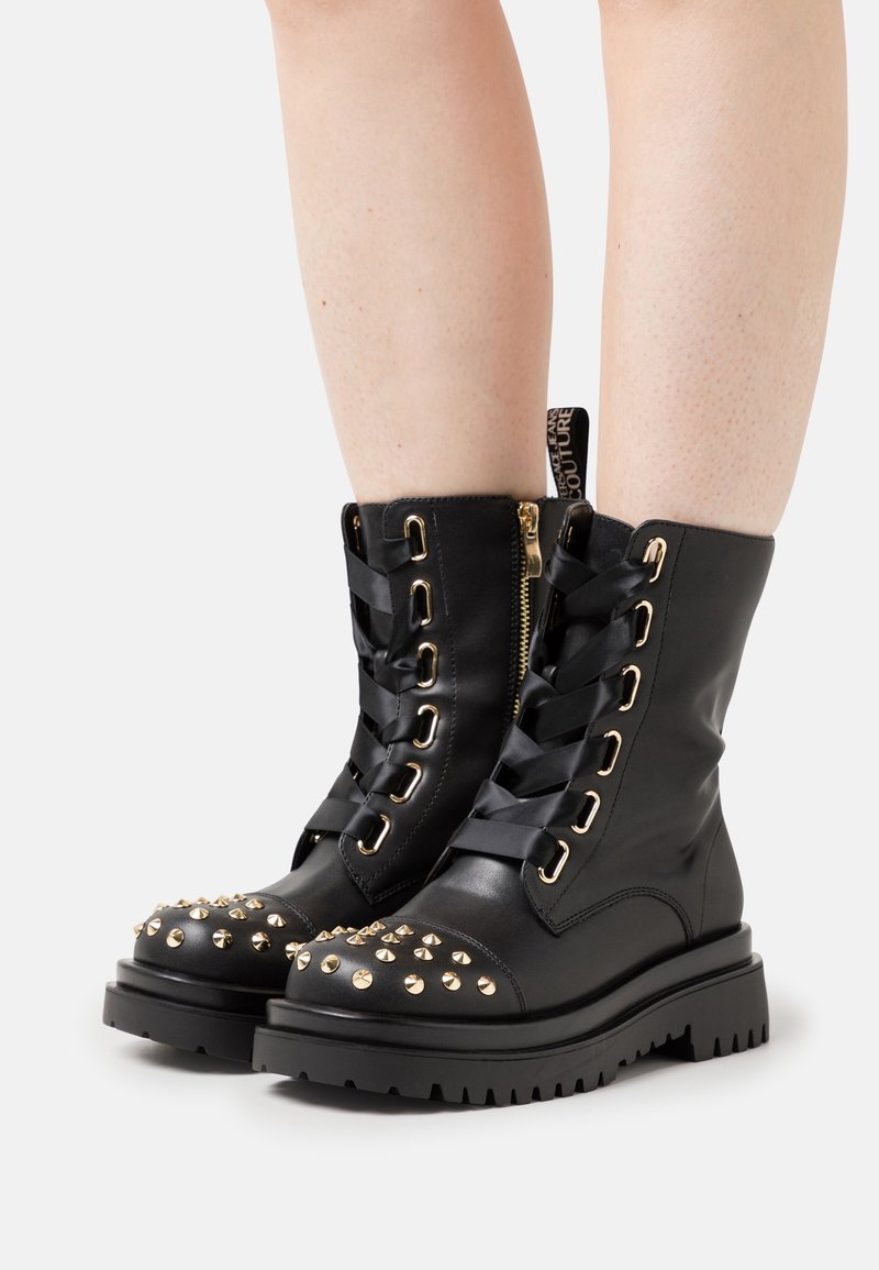 Bottes en cuir noir mi-mollet avec des clous dorés sur les orteils, des lacets larges en ruban noir, des œillets dorés, des fermetures éclair latérales et des semelles épaisses à crampons portées sur des jambes nues.