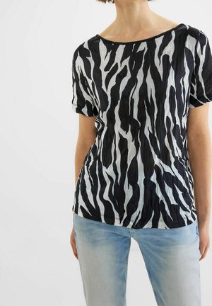 Kobieta ubrana w czarno-białą bluzkę w paski zebry z krótkim rękawem i jasnoniebieskie dżinsy, stoi na tle jednolitego białego tła.