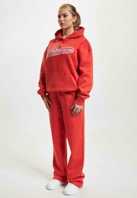 Rød hettegenser med hvit skriftlogo, frontlomme og matchende røde joggebukser, kombinert med hvite sneakers. Laget av bomull, avslappet passform.