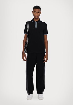 Black polo shirt with blue striped collar, paired with black pants featuring gray side panels and white sneakers. Model holds a black bag.