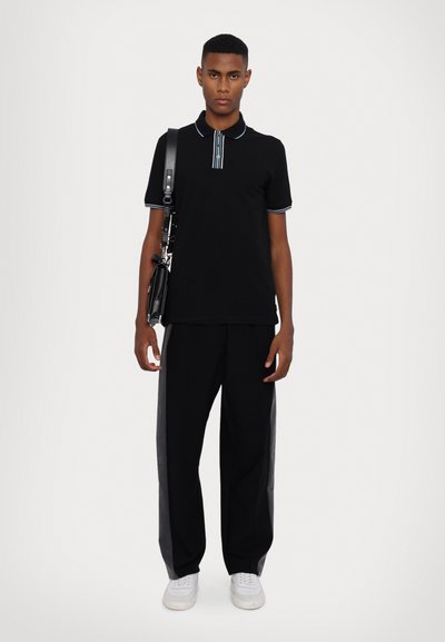 Tricou polo negru cu guler în dungi albastre, asortat cu pantaloni negri cu panouri laterale gri și adidași albi. Modelul ține o geantă neagră.