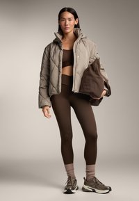 Giacca imbottita trapuntata color nocciola con colletto alto, top corto marrone e leggings marroni. Il modello indossa scarpe da ginnastica grigie con calzini rosa.