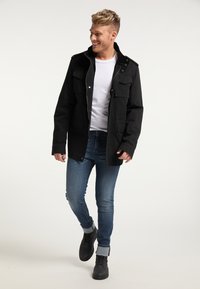 Černá bunda se čtyřmi předními kapsami, zapínání na knoflíky a zip, nošená přes bílý tričko, v kombinaci s modrými džíny a černými vysokými teniskami.