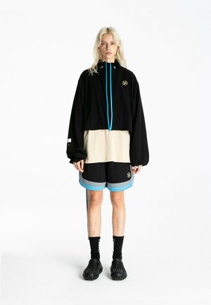 Persona de pie con una chaqueta negra extragrande con cremallera azul, camisa beige, pantalones cortos negros con ribete azul, calcetines negros y zapatos negros con textura.