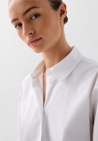 Camicia bianca con colletto classico, caratterizzata da una texture liscia e una leggera lucentezza. Presenta un design pulito e su misura.