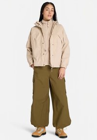 Beige oversized jas met een hoge kraag en drukknoopsluiting, gecombineerd met losse olijfgroene cargobroeken en bruine wandelschoenen.