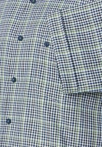 Korte mouwen bloes met een blauw, wit en groen ruitpatroon, voorzien van een borstzak en blauwe knopen. Van katoen.