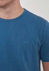 T-shirt in cotone blu con collo rotondo, con un piccolo logo ricamato nella zona sinistra del petto. Maniche corte e una texture morbida.