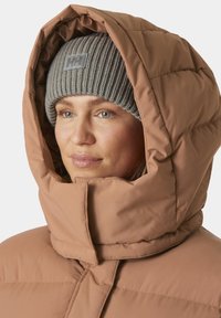 Giacca puffer in un caldo color nocciola, con colletto alto e cappuccio oversize. Abbinata a un berretto in maglia a righe grigio. Tessuto liscio e isolante.