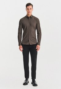 Mężczyzna stoi twarzą do przodu, ubrany w brązową koszulę w wzory z guzikiem, czarne spodnie i czarne wypolerowane buty na białym tle.