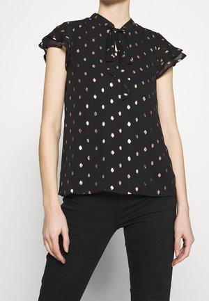Vrouw draagt een zwarte korte mouwen blouse met metallic polkadots en een strik aan de hals, gecombineerd met een zwarte broek, getoond van nek tot heupen.
