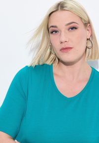 Tyrkysové tričko s výstřihem do V a krátkými rukávy, měkká textura a přiléhavý střih. Zobrazeno s hladkými, čistými liniemi a bez viditelných vzorů.