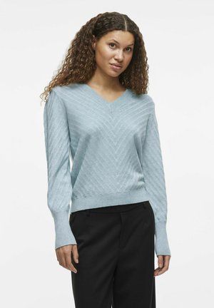 Pull en V bleu clair avec un motif en chevron texturé, des manches longues et des poignets côtelés, associé à un pantalon noir.