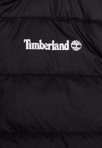 Svart pufferjakke med Timberland-logo i hvitt, laget av et glatt, vannavstøtende stoff med quiltet polstring for isolasjon.