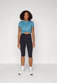 Kratka telovadna majica v barvi tirkizne z vozelom spredaj, kombinirana z visokim pasom črnimi pajkicami in belimi supergami. Enostaven dizajn z logotipi.