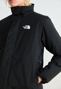Zwarte jas met hoge kraag, waterafstotende stof, een rits aan de voorkant en het The North Face-logo op de borst. Voorzien van zijzakken met een koord.