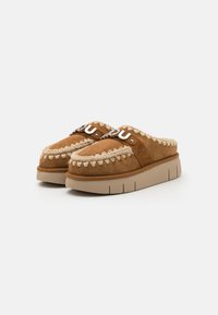 Brune semskede slip-on sko med en tykk beige såle, med dekorativ søm og et logo på stroppen.
