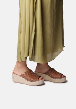 Oseba, ki nosi olivno zeleno plisirano krilo in rjave tkanine iz usnja z odprtimi prsti na platformah z juto podplati na belem ozadju.