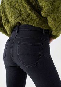 Jeans negros ajustados con bolsillos traseros, combinados con un suéter de punto verde texturizado, mostrados desde la cintura hasta la mitad del muslo.