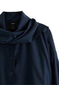 Marineblå bluse med cowl-hals, knappet front og lange ærmer. Stoffet har en glat, mat tekstur. Etiketten er synlig indeni.