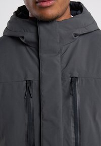 Dark gray insulated jacket with a high collar, two zippered pockets, and subtle sheen. Features minimalistic design and smooth texture.