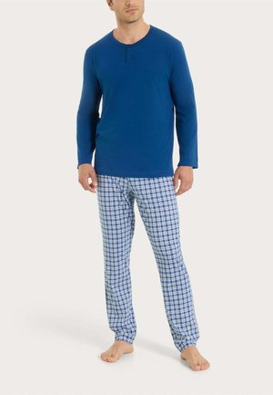 Uomo che indossa una maglia henley a maniche lunghe blu e pantaloni da pigiama a quadri blu e bianchi, in piedi scalzo su sfondo chiaro.