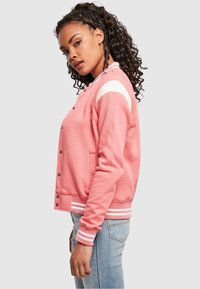 Ružičasta jakna u stilu varsity s bijelim prugama na ramenima, rebrastim manžetama, kopčama na pritisak i bočnim džepovima. Mekana tekstura tkanine.