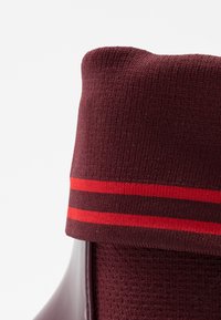 Calcetín burdeos de punto con dos rayas rojas horizontales, doblado sobre el talón brillante de un zapato oscuro, sobre fondo blanco.