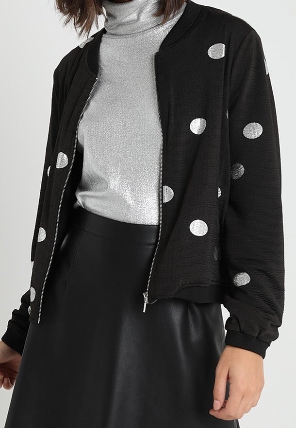 Giacca bomber nera con pois argentati, zip frontale, polsini a costine e orlo. Indossata sopra un dolcevita argentato metallico e una gonna in pelle nera.