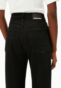 Jean en denim noir à taille haute, avec des poches arrière et un écusson en cuir noir embossé du nom "ARMEDANGELS" sur la ceinture arrière.