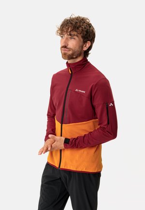 Veste zippée bordeaux et orange avec un col haut, une section inférieure texturée et une poche zippée noire sur le bras. Logo sur la poitrine.
