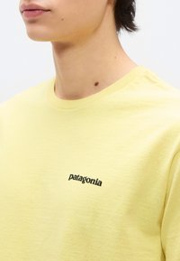 Κοντινό πλάνο στον λαιμό και τον ώμο ενός ατόμου που φοράει ανοιχτόκίτρινη μπλούζα Patagonia με λογότυπο στο στήθος, πάνω σε λευκό φόντο.