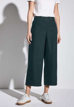 Pantaloni a gamba larga verde scuro con una texture liscia, dotati di vita alta e tre bottoni decorativi sul davanti, abbinati a sneaker bianche.