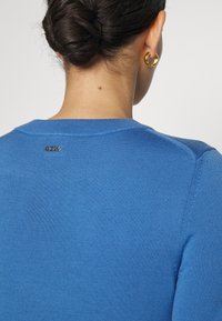 Mujer con el cabello oscuro recogido en un moño, que lleva pendientes de aro dorados y un suéter azul con un pequeño logo metálico de "BOSS" en la parte superior de la espalda.