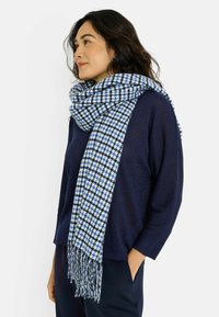 Bufanda de cuadros azules y blancos hecha de tela suave, con bordes con flecos y un diseño acogedor y en capas, llevada sobre un suéter azul marino.