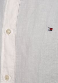 Hvid linnedskjorte med en let tekstur, der har en knaplukning og et lille broderet logo i rød og blå.
