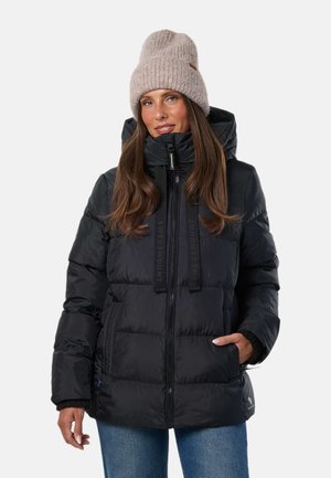 Mujer con una chaqueta acolchada negra con capucha y un gorro beige de punto, de pie con una mano en el bolsillo contra un fondo liso.