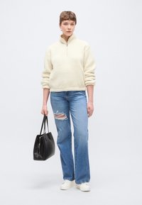 Kremasti pulover od flisa s četvrtastim patentnim zatvaračem, uparen s lagano plavim poderanim trapericama i bijelim tenisicama. Drži crnu torbu za nošenje.