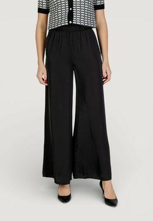 Mujer con pantalones negros de pierna ancha, tacones altos negros y una blusa de manga corta con botones y estampado, de pie frente a un fondo liso.