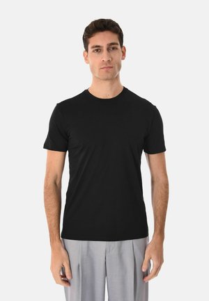 Uomo con capelli corti e scuri che indossa una maglietta nera aderente a maniche corte e pantaloni grigio chiaro, in piedi davanti a uno sfondo bianco semplice.