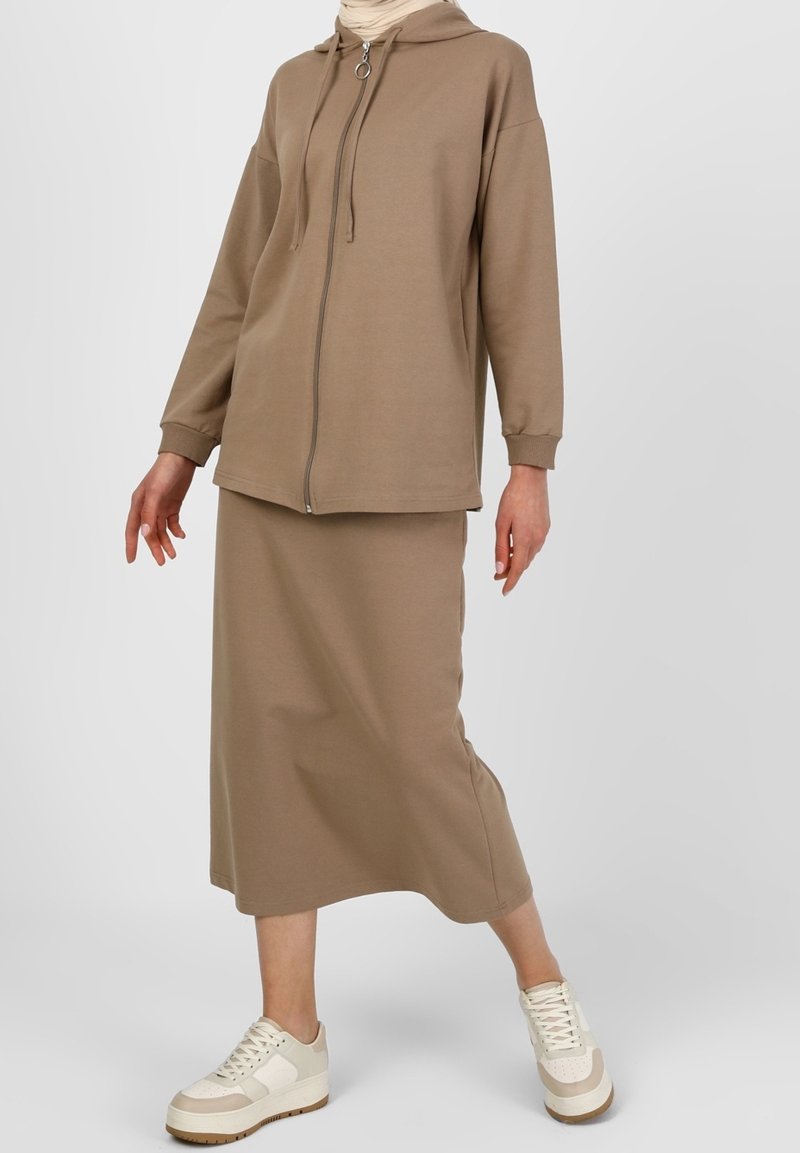 Femme portant un sweat à capuche beige zippé, une jupe midi assortie, des baskets blanc cassé et un foulard clair sur un fond uni.