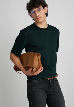 Tamnozeleni majica kratkih rukava, sivi traperice i smeša kožna torba s valjkastim vrhom, držana jednom rukom protiv jednostavne pozadine.