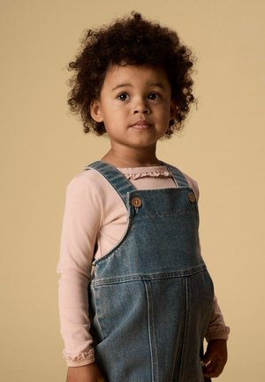 Jong kind met krullend haar, gekleed in een lichtroze longsleeve en blauwe denim tuinbroek, staand tegen een beige achtergrond.