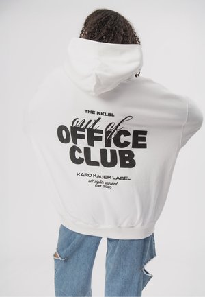 Person mit lockigem Haar, die einen weißen Hoodie mit dem Text "out of OFFICE CLUB" auf dem Rücken und zerrissene blaue Jeans trägt.