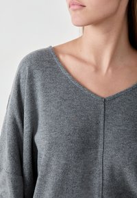 Grau gestrickter Pullover mit V-Ausschnitt, der eine weiche Textur, eine lockere Passform und abgesenkte Schulternähte aufweist. Minimalistisches Design, keine sichtbaren Muster.