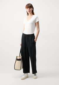 Weißes V-Ausschnitt-T-Shirt, lockere schwarze Hose mit elastischem Bund, weiße Sneakers und eine beigen Tragetasche mit einem schwarzen Akzent.