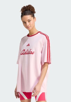 Jeune femme aux cheveux bouclés porte un T-shirt rose oversize Adidas avec des rayures rouges et un short assorti rouge et blanc, debout devant un fond clair.