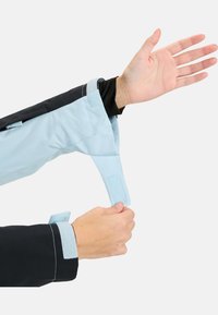 Jasnoniebieska wodoodporna kurtka z regulowanym mankietem. Czarna tkanina na ramieniu. Widoczna ręka chwycająca pasek zapięcia mankietu.